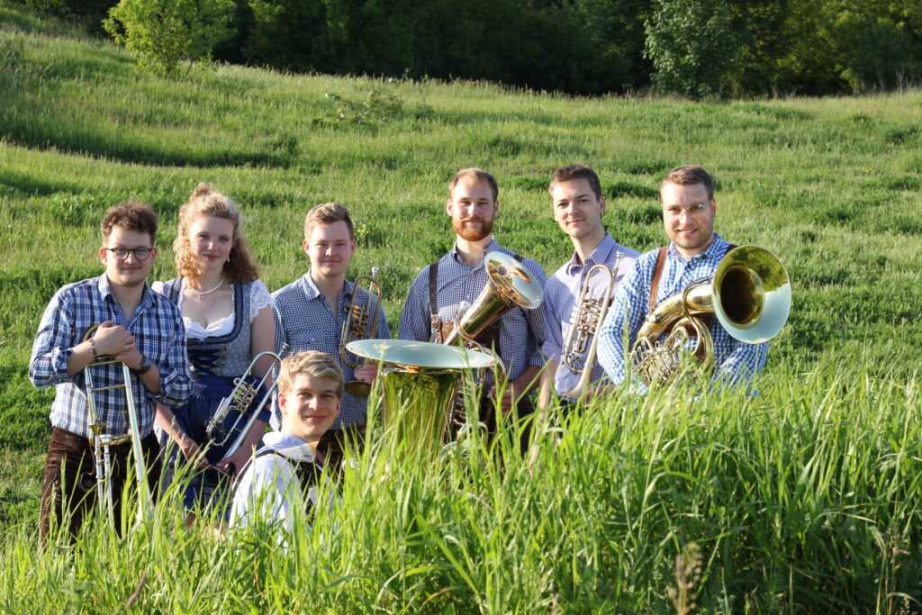 Die Band in Tracht steht im hohen Gras einer sonnigen Wiese, Posaune, Flügelhörner, Tenorhorn und Tuba im Arm.
