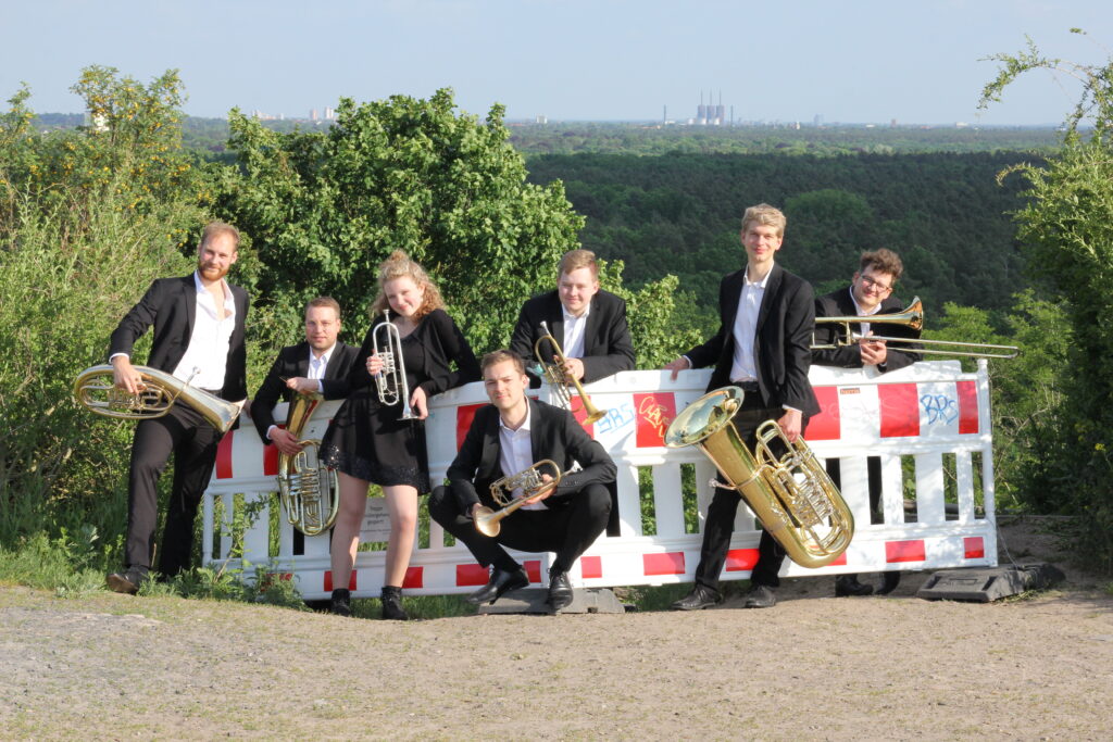 Die Band im schwarzen Anzug auf einer sandigen Anhöhe, angelehnt an eine rot-weisse Absperrbake, im Hintergrund Berliner Wald und die Schornsteine eines Kraftwerks.