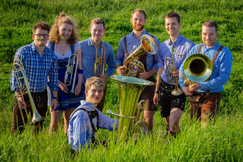 Gruppenbild des Ensembles in Tracht auf einer Wiese, mit Trompeten, Posaunen, Tenorhorn und Tuba — jede und jeder mit einem Grashalm zwischen den Zähnen.