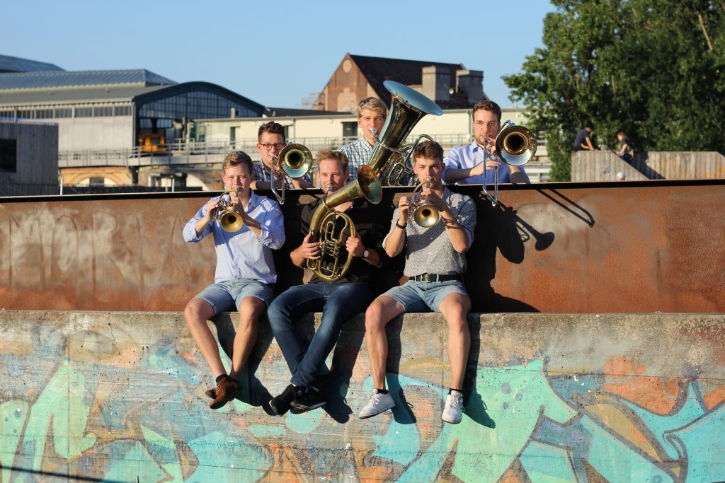 Die Band spielt sitzend auf einer mit Graffiti besprühten Mauer, im Hintergrund Berliner Hallendächer.