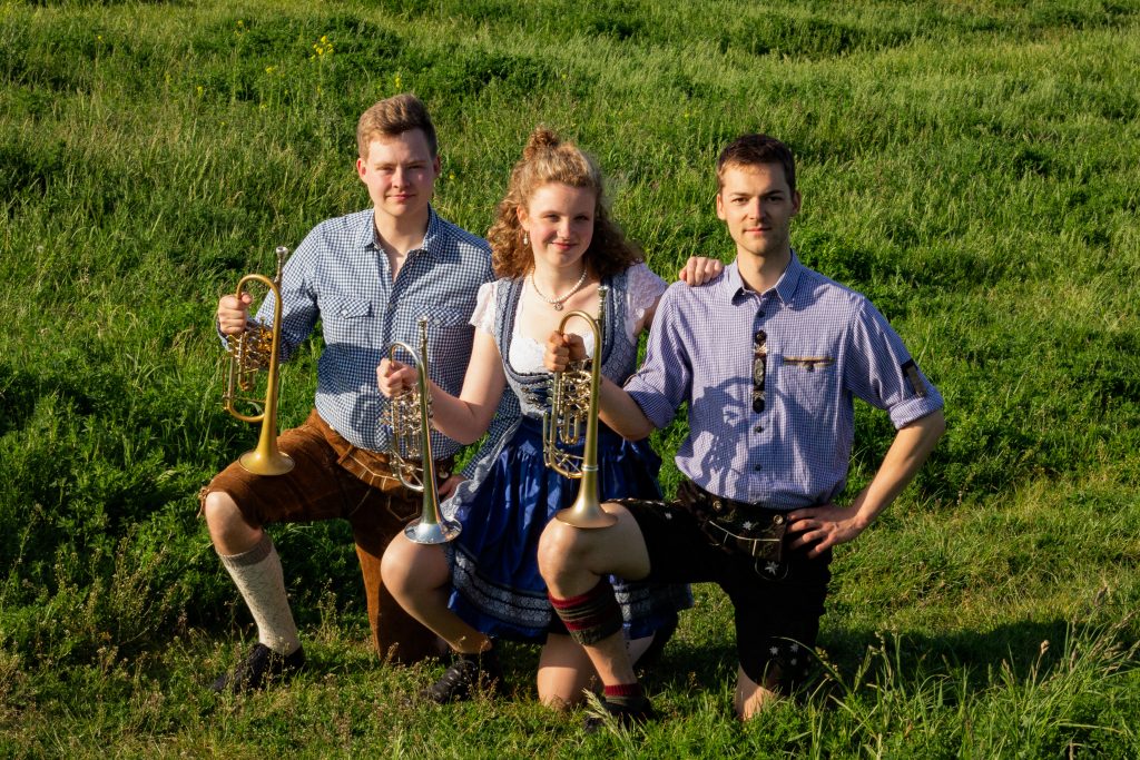 Drei Mitglieder von SpreeBlech in Tracht knien mit ihren Trompeten auf einer sonnigen Sommerwiese.